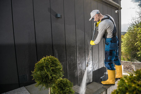 Male Caucasian Home Owner at His 40s Wearing His Work Clothes Cleaning His Yard By Washing the Walls of the House and Nearby Concrete Slabs with Pressure Washer.の写真素材