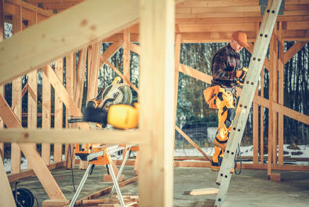 Caucasian Worker Going Up the Stepladder to Check the Roof Section of Wooden Skeleton of the Residential House Under Construction.の写真素材