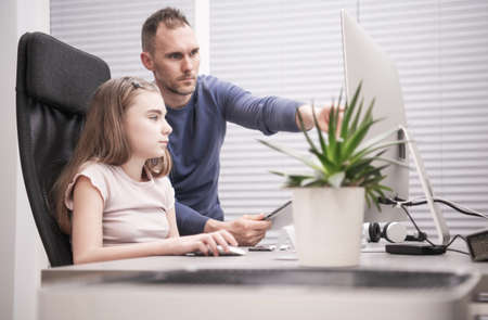 Caucasian Family of Father and Daughter Sitting in Front of the Computer Looking at the Monitor. Proud Father Instructing His Young Daughter in the Use of Computer Programs Useful During Her School Studies.の写真素材