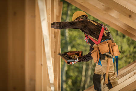 Caucasian Male Worker Wearing Safety Harness and Hard Hat on the Construction Site of Wooden Residential Building. Working in the Roof Section, Driving Nails Into Wood Planks Using Nail Gun.の写真素材