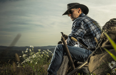 American Cowboy in His 40s Enjoying Free Time on His Countryside Ranch. Seating on a Piece of Wooden...