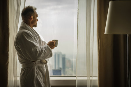 Caucasian Man Getting Ready For a New Day While in Business Travel. Looking Through the Hotel Room Window and Drinking Coffeeの写真素材