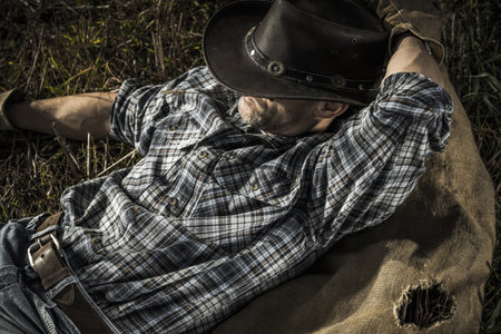 Caucasian American Cowboy Resting on a Ground and Chew on Straw. Taking Short Break on a Farm.の写真素材