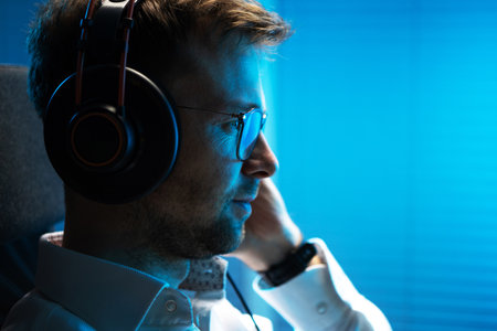 Caucasian Man in His 30s Listening Podcast on His Computer Using Professional Wireless Headphones. Dark Blue Illumination Scene.の写真素材