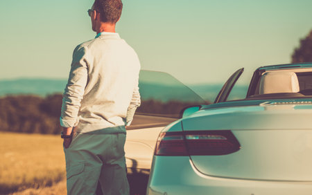 Wealthy Caucasian Man Standing Backwards Next to His Modern White Convertible Car Enjoying Peaceful Sunset. Happy Life.の写真素材