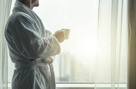 Caucasian Man in His 40s with Coffee Mug in His Hands Taking a Look From His Apartment Windowの写真素材