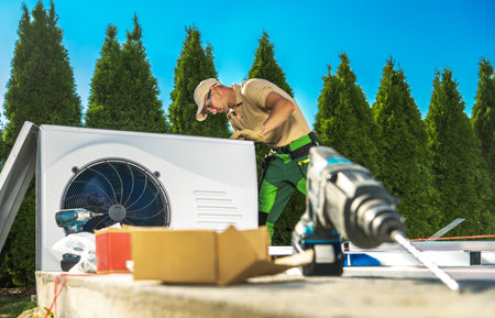 HVAC technician wearing a hat and green pants is diligently working on an air conditioner or heat pump unit outdoors.の写真素材