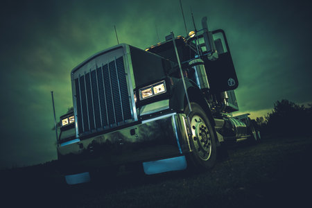 A close-up image of a semi-truck parked on a dark road at night. The truck is illuminated by its...