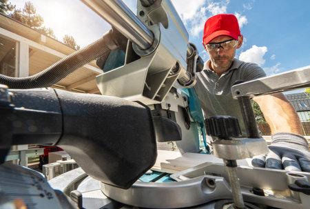 A construction worker wearing safety glasses and a red cap uses a miter saw to cut a piece of wood...