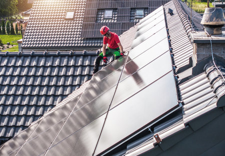 A worker wearing a red hard hat and green pants installs solar panels on a residential roof. The...