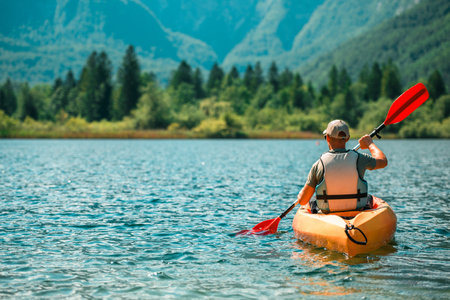 A man kayaking on a calm lake, with a backdrop of lush green trees and a distant mountain range. The water is clear and blue, reflecting the sunny sky. He is wearing a life jacket and using a red paddle to propel himself forward.の写真素材