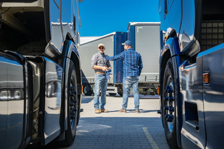 Two truck drivers engage in a conversation about logistics while standing between parked trucks in a transport yard. The bright blue sky adds to the vibrant atmosphere of this bustling environment.の写真素材