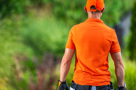 A gardener dressed in an orange shirt stands with his back facing the camera, holding gardening...