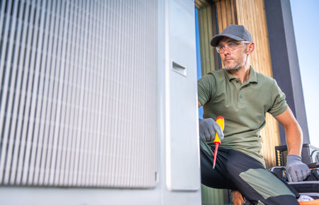 A technician in a green polo shirt and cap performs maintenance on an air conditioning unit. He uses a screwdriver and looks focused while working outdoors on a sunny day, surrounded by wooden walls.の写真素材