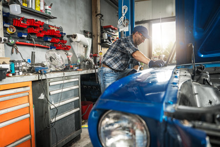A skilled mechanic is focused on repairing a classic blue car in a well-organized garage workshop. Tools and equipment surround him as he assesses the vehicle's condition, ensuring a meticulous restoration process.の写真素材