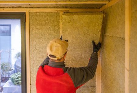 A construction worker wearing gloves and a cap carefully installs insulation panels in a modern home's interior.の写真素材