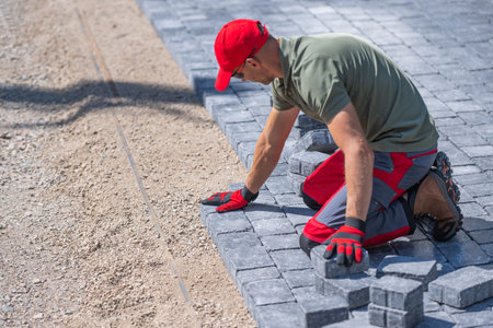 A worker is carefully laying down pavers on a prepared gravel base, focusing on alignment and...