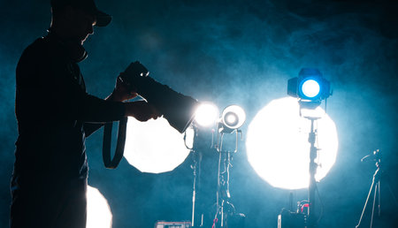 A photographer in silhouette adjusts equipment amid bright lights and smoke during a nighttime studio session.の写真素材
