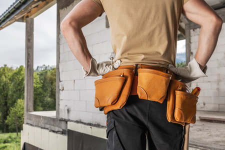 A construction worker stands with hands on hips, wearing a tool belt filled with equipment at a building site.の写真素材