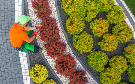 A gardener in bright attire is pruning vibrant shrubs in a meticulously designed garden surrounded...
