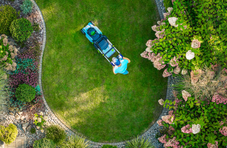 A person is mowing a perfectly shaped circular lawn in a vibrant garden filled with lush green plants and blooming flowers on a sunny day.の写真素材