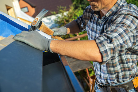 A skilled worker is installing roofing materials on a home's upper section, using a hammer while wearing gloves. The sun shines brightly above, highlighting his focused effort.の写真素材