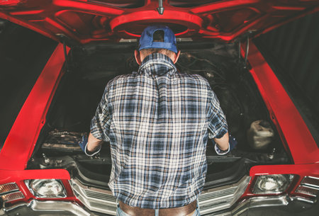 A dedicated car enthusiast works on a classic vehicle in a garage, focusing on the engine under the hood with tools nearby.の写真素材