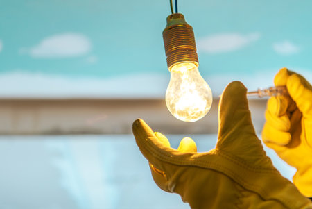 A person wearing yellow gloves holds a lit light bulb, adjusting it as bright light emits in a workspace with a blue background.の写真素材