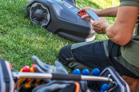 A person is using a smartphone to operate a robotic lawn mower on a well-maintained lawn, with landscaping tools visible nearby.の写真素材