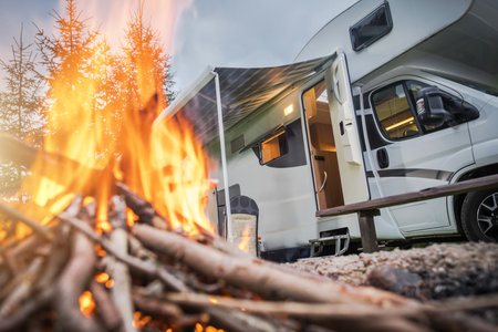 A warm fire crackles in front of a cozy RV, set amidst trees as the sun sets. The inviting atmosphere highlights nature and relaxation in the great outdoors.の写真素材
