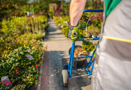 A gardener is pushing a cart filled with plants in a lush nursery, surrounded by a variety of colorful flowers and greenery on a bright day.の写真素材