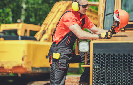A construction worker in orange gear inspects machinery at a busy site, ensuring safety and proper operation with protective equipment.の写真素材