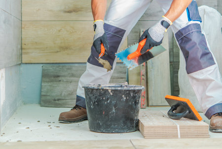 A worker focused on laying tiles, using tools next to a bucket of adhesive and tile pieces in a contemporary setting.の写真素材