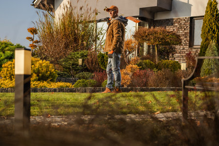 In a well-kept garden, a man stands confidently with landscaping equipment, surrounded by colorful plants and trees, basking in the warm autumn light.の写真素材