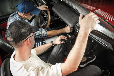 Two mechanics work together on a classic car's interior, focused on repairs inside a garage...