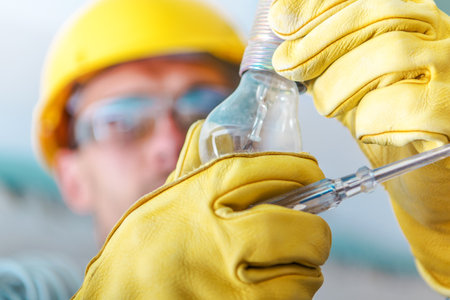 A worker in a yellow hard hat and gloves carefully replaces a light bulb in an industrial...