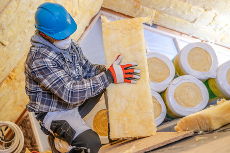 A construction worker is carefully installing insulation materials in a residential attic, wearing...