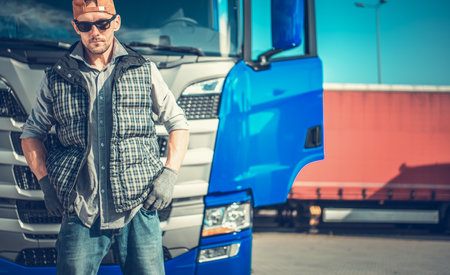 A man dressed in work attire poses with confidence near a blue truck, reflecting hard work and dedication in a transport setting during daylight.の写真素材