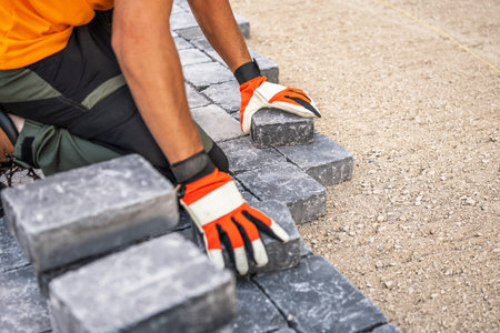 A construction worker is carefully laying gray stone blocks on a prepared surface. The worker, wearing an orange shirt and gloves, focuses on aligning the blocks for a new outdoor pathway during daylight hours.の写真素材