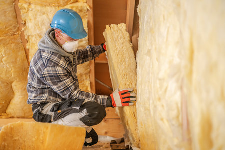 A worker wearing a blue hard hat and safety gloves is carefully placing insulation material against a wall. The setting is an unfinished building, highlighting construction efforts and safety measures in progress.の写真素材