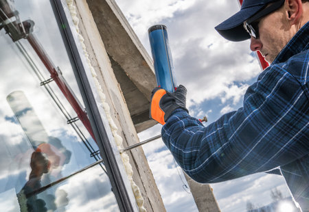 A construction worker applies sealant to install glass in a building while using a caulking gun. The cloudy sky serves as a backdrop, highlighting the ongoing construction activity during the day.の写真素材