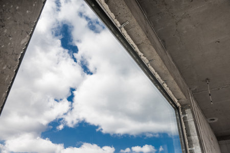 A large window frames a view of the sky filled with fluffy clouds. The scene captures the contrast between the raw concrete of the unfinished building and the vibrant blue sky beyond, highlighting both nature and construction.の写真素材