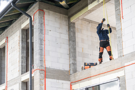A worker in safety gear carefully measures the height of a wall framework at a construction site. Bright orange electrical wires are visible on the walls, indicating ongoing electrical installation work.の写真素材