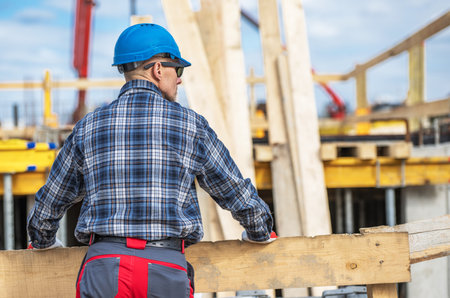 A construction worker wearing a blue hard hat and plaid shirt observes the ongoing work at a construction site. Wooden beams and heavy machinery are visible in the background under a bright sky.の写真素材