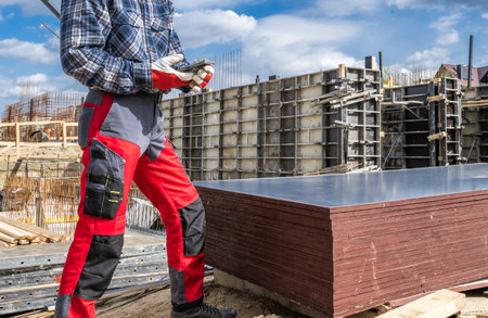 A construction worker wearing a checkered shirt and red pants is checking equipment while standing beside stacked plywood at a busy construction site. Steel framework and scaffolding are visible in the background under a blue sky.の写真素材