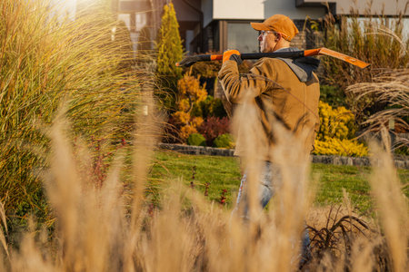 A gardener in a brown jacket and orange cap carries pruning tools while carefully navigating through tall grasses in a beautifully landscaped backyard, illuminated by warm sunlight at sunset.の写真素材