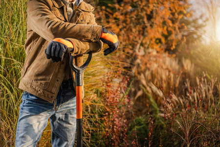 A man dressed in a brown jacket and gloves rests on his shovel while standing among colorful autumn foliage. The warm sunlight illuminates the plants around him, indicating a tranquil gardening moment.の写真素材