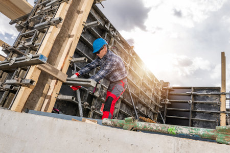 A construction worker in a blue helmet prepares concrete formwork at a building site during the day. Heavy wooden beams and steel supports are used to ensure stability as clouds partially obscure the sun.の写真素材