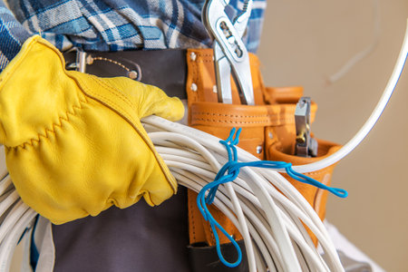 In a well-lit space, an electrician secures a bundle of cables with a blue tie while wearing a yellow glove. The professional is equipped with various tools, highlighting the meticulous preparation for electrical installation.の写真素材