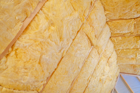Bright yellow insulation material fills the ceiling of a residential attic. Workers prepare the area for better energy efficiency, ensuring each section is securely placed. The installation process occurs during daylight hours.の写真素材